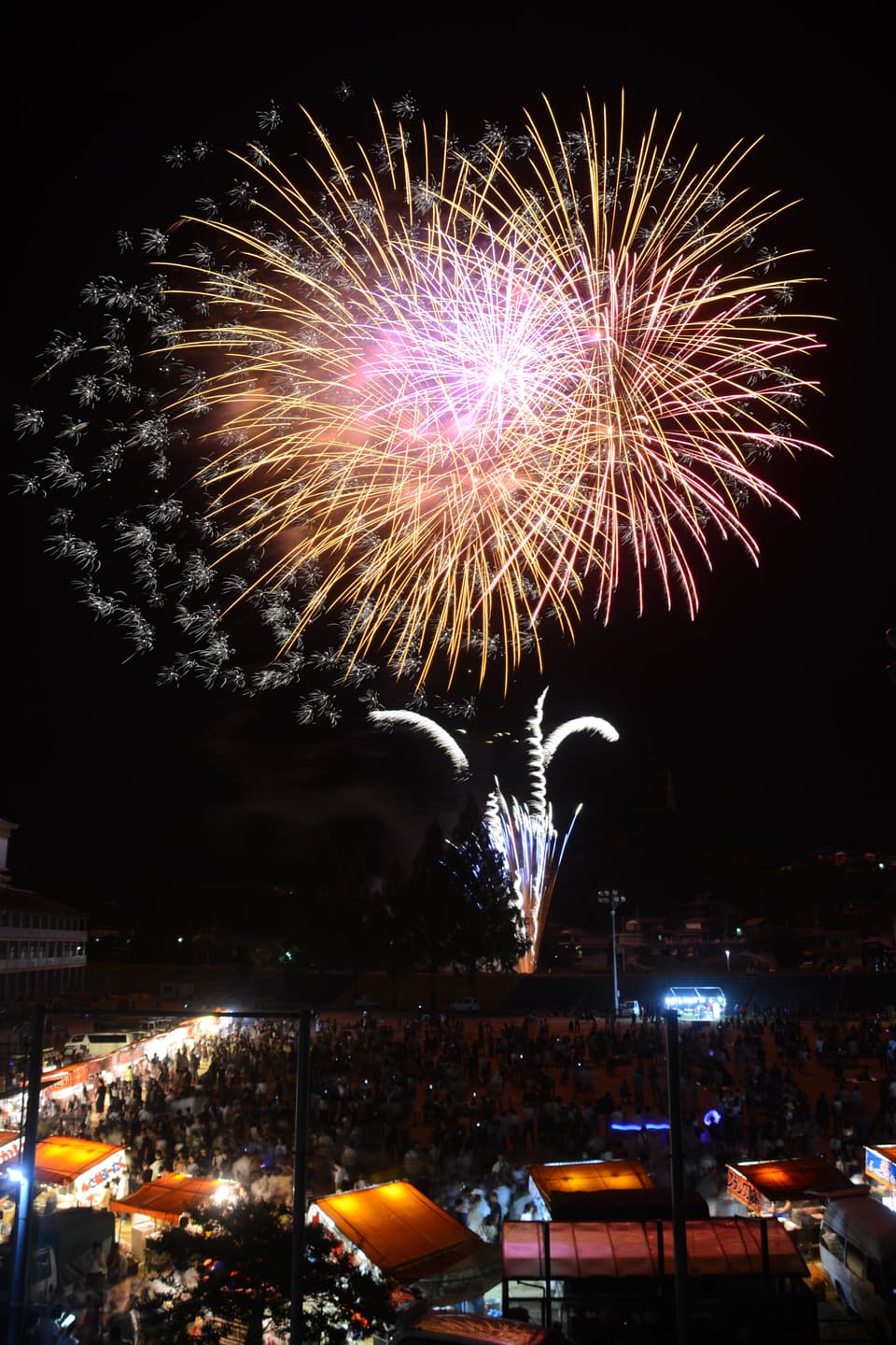 つるぎ町夏祭りで夜空に花火が上がる様子、縦型写真、赤系の大きな花火が写真の４分の３を占める、写真３分の１は貞光中学校のグラウンドに屋台が並び多くの人が集まる様子が写っている
