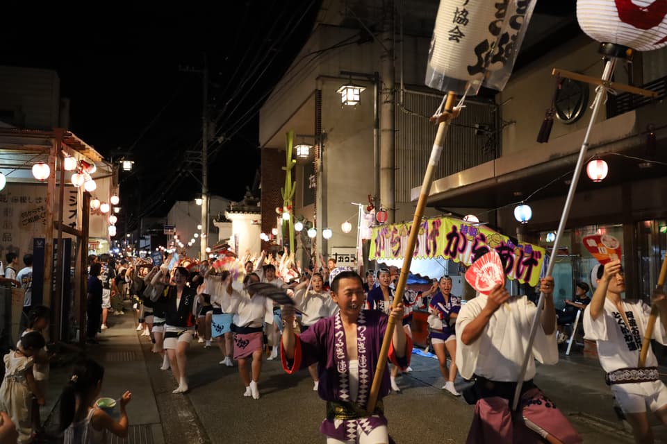 貞光2層うだつの町並みをいく阿波踊りの連。前方に提灯をかかげた人3人、その後を５列ほどに並んで大勢の人の踊りが続く。道の両側には観客がおり、からあげの屋台も出ている。